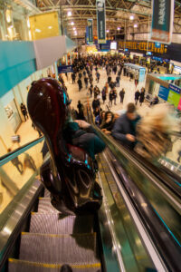 Cellist with their cello case riding down an escalator into the busy concourse of Waterloo station, London.