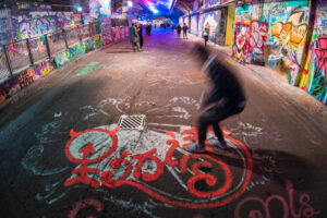 Long-exposure photograph in London's Leake Street graffiti tunnel — graffiti-covered walls, a skateboarder mid-motion blur centred over a tag on the floor.