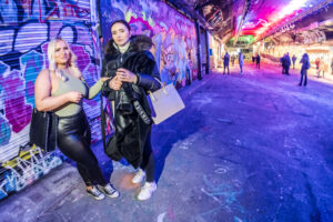 Two young women posing against vibrant graffiti walls in the Leake Street tunnel under Waterloo, with red and blue light bouncing off the floor.