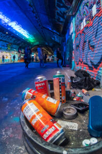 Close-up of artists' spray-paint cans on a drum in the Leake Street tunnel under Waterloo, with figures walking the graffiti corridor behind.