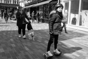 Black-and-white street scene of a girl in a helmet riding a hoverboard past a woman walking a small dog, on a Kingston high street.