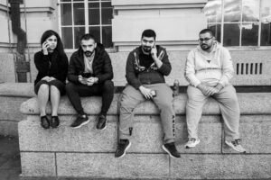 Black-and-white candid of four young men on phones sitting on a stone bench outside a city building, end-of-day light.
