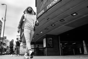 Black-and-white low-angle street portrait of a man in a tracksuit walking past the entrance of Wood Green tube station, London.