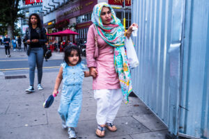 Colour candid street scene of a mother in a pink and turquoise headscarf walking with her young daughter on the Wood Green high street at dusk.