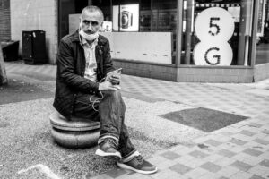 Black-and-white street photograph of an older man sitting on a stone bollard with earphones in, beside a shop window with a stylised "5G" sign, Wood Green.