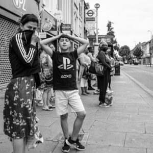 Black-and-white candid of a boy striking a mock-shocked pose with hands on his head and tongue out, his mother behind hiding her face, at a Wood Green bus stop.