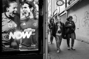 Black-and-white candid of three young women walking past a large football-themed advertising poster on a Wood Green street.