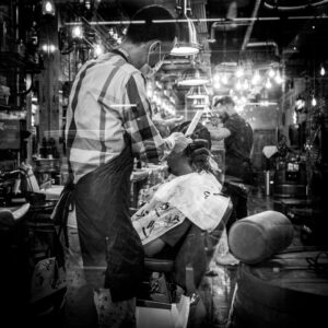 Black-and-white street photograph through a Wood Green barber's window: a barber in a checked shirt and mask cuts a child's hair, the room layered with reflections through the glass.