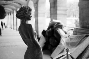 Black-and-white photograph of a bronze sculpture of a woman in a flowing dress in the arcades of Place des Vosges, Paris.