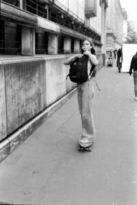 Black-and-white candid of a young woman skateboarding down a Paris pavement past a classical stone building, looking over her shoulder.