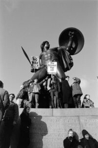Documentary photograph of demonstrators at the 1991 Gulf War anti-war protest in London, holding "No War" and "No Blood for Oil" placards in front of a heroic statue.