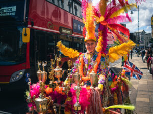 Colour candid of a man in costume cycling through Wimbledon, vivid yellow and orange feathered headdress catching the sun, a London bus passing behind.