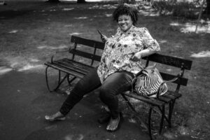 Black-and-white candid of a woman laughing on a park bench, barefoot with a striped handbag beside her, dappled summer light.
