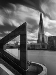 Black-and-white long-exposure photograph of The Shard rising through a triangular concrete sculpture on the South Bank, streaked cirrus clouds.