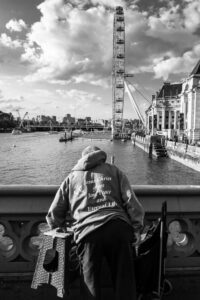 Black-and-white photograph of a person on Westminster Bridge wearing a hoodie printed "Jesus Christ gives Joy, Peace and Eternal Life", looking out toward the London Eye.