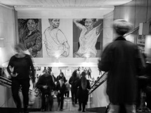 Black-and-white photograph of commuters moving below the three large painted mural portraits at Brixton Underground station, motion blur on the figures.