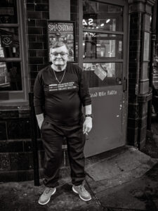 Black-and-white street portrait of an older person in glasses standing outside an Irish pub in Brixton, a Splashdown poster in the window and "Cead Mile Failte" painted on the door frame.