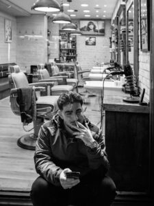 Black-and-white candid of a young man sitting on the step of a Brixton barber shop, smoking and scrolling his phone, the empty chairs of the shop behind him.