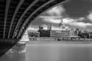 Long-exposure black-and-white photograph looking under the arches of Blackfriars Bridge toward St Paul's Cathedral and the City of London skyline.