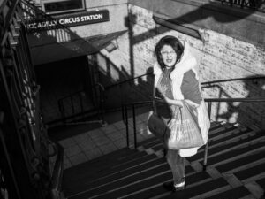 Black-and-white candid of a woman emerging from the steps of Piccadilly Circus tube station carrying shopping bags, shadow of the station sign on the wall behind.