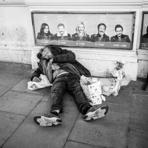Black-and-white documentary photograph of a person sleeping rough on the pavement at Piccadilly Circus beneath a row of West End theatre posters of performers.