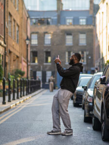 Colour street photograph of a man pausing in the middle of a side street near Brick Lane to take a photograph with his phone, parked cars and Georgian terraces around him.