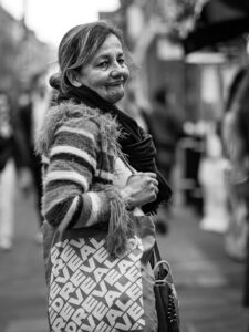 Black-and-white candid of an older woman with a fringed striped jumper carrying a "REVEAL"-printed tote bag, half-smiling on a London street.