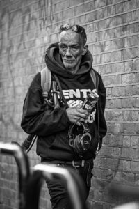 Black-and-white street portrait of an older man with sunglasses on his forehead, a hoodie and headphones around his neck, standing against a brick wall holding a can.