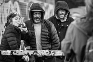 Black-and-white candid of three older friends in hooded jackets at a London street market, the woman on the left vaping or smoking.