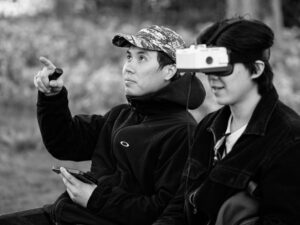 Colour candid of two young men in a park, one wearing a vintage stereo viewer/camera as a headset while pointing into the distance, the other holding a phone.