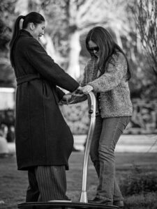 Black-and-white candid of two women playing on a children's playground roundabout in a London park, dappled afternoon light.